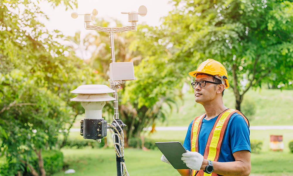 A person wearing a hard hat and safety vest holds a tablet while inspecting weather monitoring equipment outdoors in a green, park-like area.