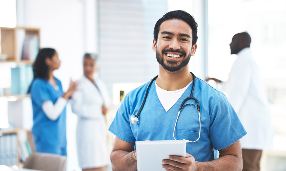 Smiling healthcare professional in blue scrubs holds a digital tablet, with a stethoscope around his neck. In the background, three colleagues in white coats are engaged in conversation.