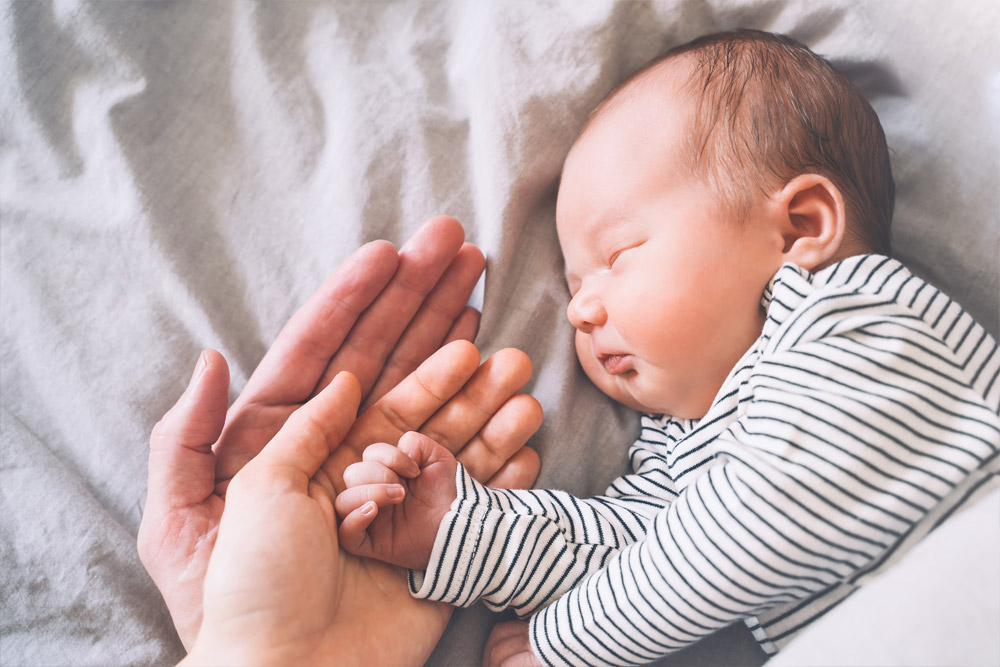 A newborn baby in a striped onesie sleeps while holding an adult’s finger. Two adult hands gently cradle the baby's hand, all resting on a soft, light-colored blanket.