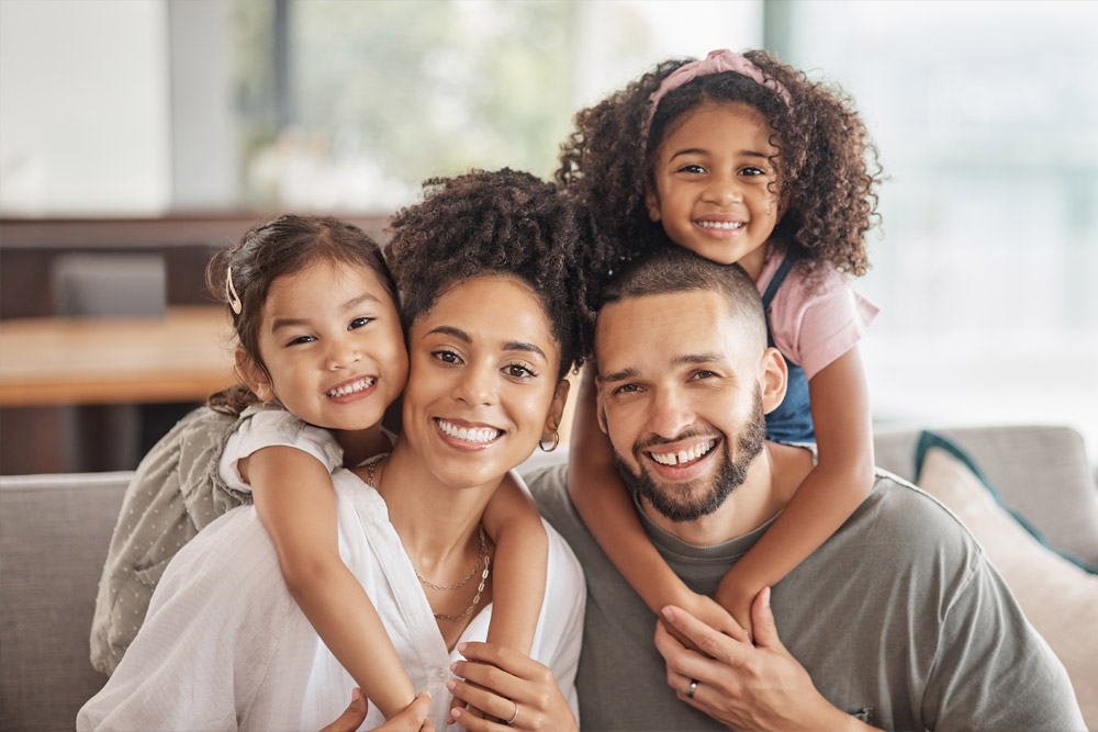 A smiling family of four poses indoors on a couch, with two young daughters hugging their parents from behind. Everyone looks happy and close together.